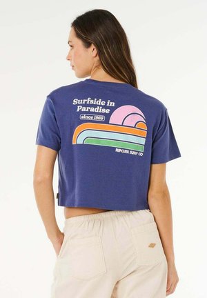 Mujer con camiseta corta azul marino con gráfico retro y texto "Surfside en el Paraíso desde 1969" y pantalones beige, de pie de perfil.