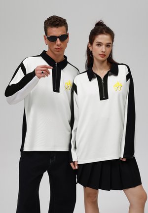 Polo unisexe à manches longues en tissu blanc et noir avec une fermeture éclair mi-longue, ornée d'un graphique de flamme jaune et de panneaux contrastés. Pantalons et jupe noirs.