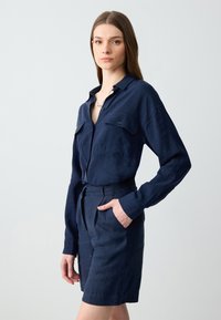 Camicia a maniche lunghe blu navy con bottoni e due tasche sul petto, abbinata a pantaloni corti coordinati. Tessuto simile al lino con una vestibilità rilassata.
