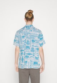 Camicia a maniche corte in tessuto blu chiaro con un motivo ripetuto di pubblicità vintage Levi's in blu scuro. Il design del colletto è classico.