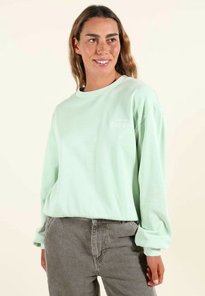 Femme souriante portant une chemise à manches longues vert clair et un jean gris, debout avec une main dans la poche devant un fond uni.