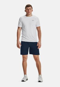 Camiseta atlética blanca de manga corta con un logo gris, combinada con shorts de color azul marino. El calzado es zapatillas deportivas de color gris claro.