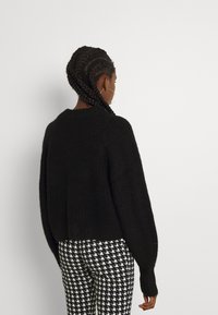 Woman with braided hair wearing large hoop earrings, a black textured sweater, and black-and-white houndstooth pants, standing against white wall.