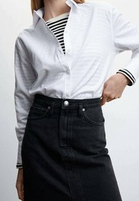 Chemise blanche à boutons avec une texture subtile sur un haut rayé noir et blanc, associée à une jupe en denim noir taille haute avec une fente à l'avant.