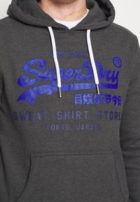 Felpa grigia con cordini bianchi. Presenta una stampa audace blu con la scritta "Vintage SuperDry" e un testo aggiuntivo in blu navy. Texture morbida.