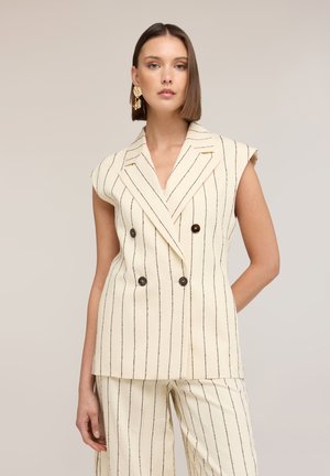 Kvinde iført hvid ærmeløs dragt med sorte, lodrette striber og dobbeltradede knapper, kombineret med matchende stribede bukser og guldringe.