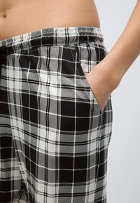 Svartvita rutiga byxor med elastisk midjeresår, använda av en person med handen avslappnat i sidofickan mot en enfärgad bakgrund.