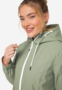 Svetle olivově zelená bunda s kapucí, předním zipem, šňůrkami a elastickými manžetami, s hladkou texturou. Mezi výrazné rysy patří kontrastní vnitřní podšívka.