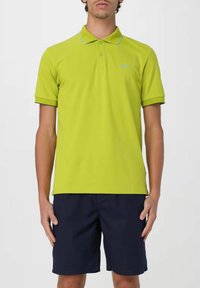 Polo shirt verde lime con colletto, cuciture azzurre a contrasto e maniche corte, abbinato a pantaloni corti blu; texture del tessuto liscia visibile.