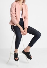 Blazer rose texturé, haut blanc, jean skinny foncé avec des bandes latérales, et chaussures noires à enfiler avec une semelle épaisse. Assis sur un tabouret blanc.