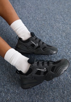 Personne portant des baskets Nike noires et grises avec des logos blancs en forme de swoosh et des chaussettes blanches côtelées sur un tapis bleu texturé.