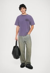 Violet medvilninis trumpas rankovės marškinėliai su juodu logotipu. Dėvimi su nudėvėtais žaliais džinsais ir juodais slip-on batais, laikant juodą maišą.