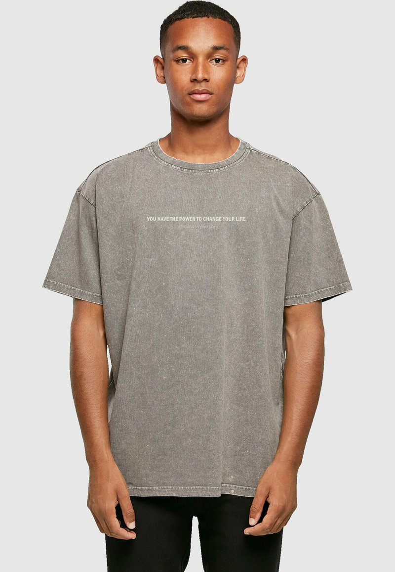 Grijze oversized T-shirt van katoen, met een ronde hals en korte mouwen, met de tekst "JE HEBT DE KRAFT OM JOUW LEVEN TE VERANDEREN" gedrukt over de borst.
