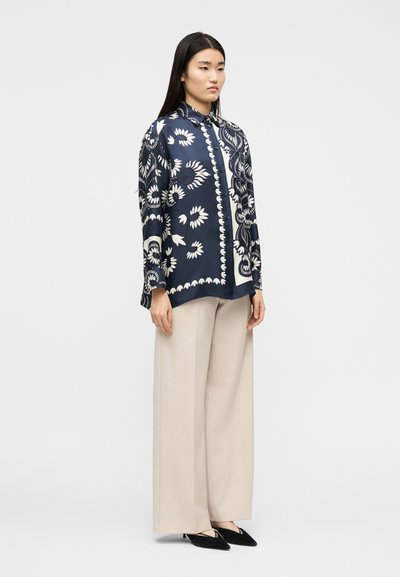 Γυναίκα που φοράει μπλε σκούρη μπλούζα με μακριά μανίκια και σχέδια, μπεζ παντελόνι με φαρδιά μπατζάκια και μαύρες μυτερές γόβες, στέκεται μπροστά σε απλό φόντο.