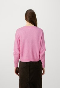 Kvinna med långt brunt hår som bär en rosa långärmad tröja och en mörkbrun denimkjol, stående vänd bort från kameran mot en enfärgad bakgrund.