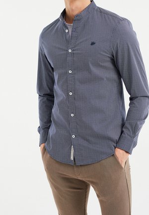 Man draagt een blauw geruit overhem met mandarijnkraag en bruine broek, handen in de zakken, tegen een effen witte achtergrond.