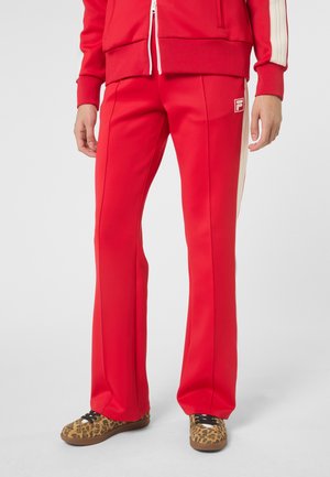 Osoba ubrana w czerwone spodnie dresowe z białymi paskami po bokach i logo, nosząca buty sportowe w panterkę na jednolitym tle.