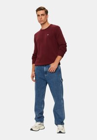 Maglione a maglia color mogano con polsini a coste, abbinato a jeans di denim blu chiaro e sneakers bianche con dettagli neri, in posa in piedi.