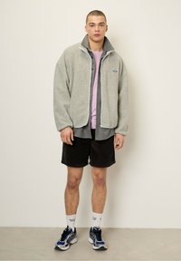 Hellgraue Fleecejacke mit hohem Kragen, über einem rosa Shirt getragen, kombiniert mit schwarzen Shorts und blauen Sneakers. Besitzt ein kleines Logo-Patch.