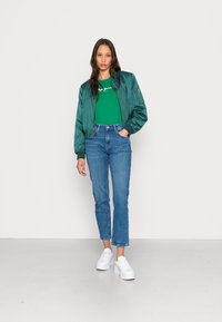 Casaco bomber verde de cetim, t-shirt gráfica branca, jeans curtos azuis e ténis de plataforma brancos. O casaco apresenta punhos canelados e detalhes de fecho éclair.