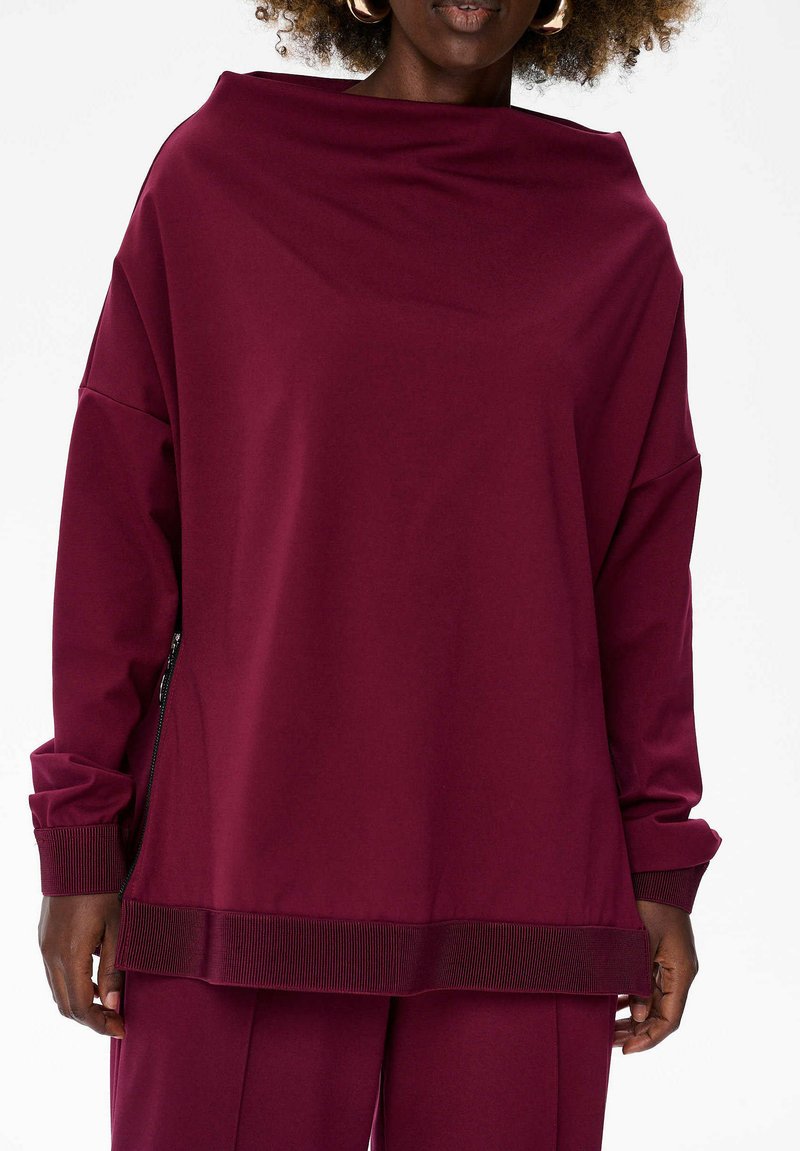 Vrouw met een oversized, langemouwen, bordeauxrode top met ribbelmanchetten en -zoom, gecombineerd met bijpassende losse broek tegen een effen achtergrond.