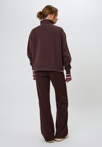 Pullover oversize marrone con colletto alto, abbinato a pantaloni svasati coordinati. Accenti a righe visibili sui polsini delle maniche. Tessuto morbido.