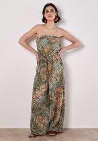 Grüner Jumpsuit mit Palmenblattmuster, trägerlosem Oberteil, tailliertem Bund, weitem Bein und leichtem Stoff. Tanfarbene Sandalen ergänzen das Outfit.