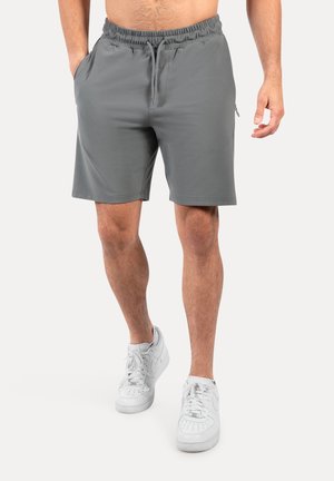 Man die grijze trekkoordshorts met zij- en achterzakken en witte sneakers draagt, staat tegen een effen achtergrond.
