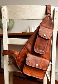 Hnědá kožená taška přes rameno se třemi předními kapsami, zaobleným dnem a bronzovými kovovými doplňky; hladká textura s mírnými záhyby.