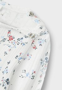 Tissu blanc à motifs floraux avec de petites fleurs bleues et rouges. Présente une texture ribbed et des pressions en métal argenté au niveau du col.
