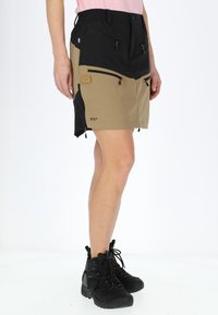 Knee-length outdoor skirt in tan and black fabric, with multiple zippered pockets and adjustable waist, paired with black hiking boots.