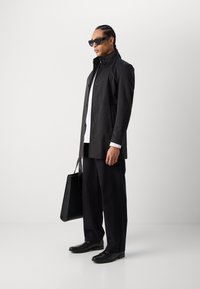 Zwarte trenchcoat met gestructureerde kraag, knoopdetails, gecombineerd met zwarte broeken en leren schoenen, met een zwarte tote bag.