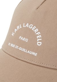 Kreminė kepurė su struktūrizuotu dizainu, su baltu tekstu: "KARL LAGERFELD PARIS" ir adresu "21 RUE ST-GUILLAUME." Medžiaga yra glotni.