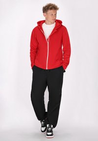 Rode zip-up hoodie met witte rits, zwarte loszittende broek en zwarte sneakers met witte en rode accenten. Model staat casual.