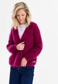 Kobieta o blond włosach, ubrana w głęboko różowy, puszysty kardigan na białej koszuli i niebieskich dżinsach, poprawiająca kardigan rękami.