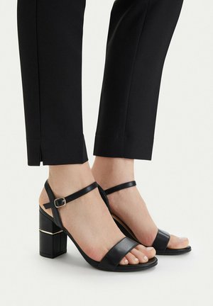 Piedi con sandali neri in pelle con tacco a blocco, cinturini alla caviglia e fibbia dorata, abbinati a pantaloni neri sartoriali con spacchi alle caviglie.