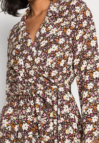 Vestido envolvente floral que presenta una mezcla de flores blancas, rosas y naranjas sobre un fondo negro, con cintura anudada y mangas largas.