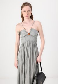 Abito maxi grigio chiaro con design plissettato, spalline sottili e dettaglio annodato sul petto. Modella che tiene in mano una borsa nera.