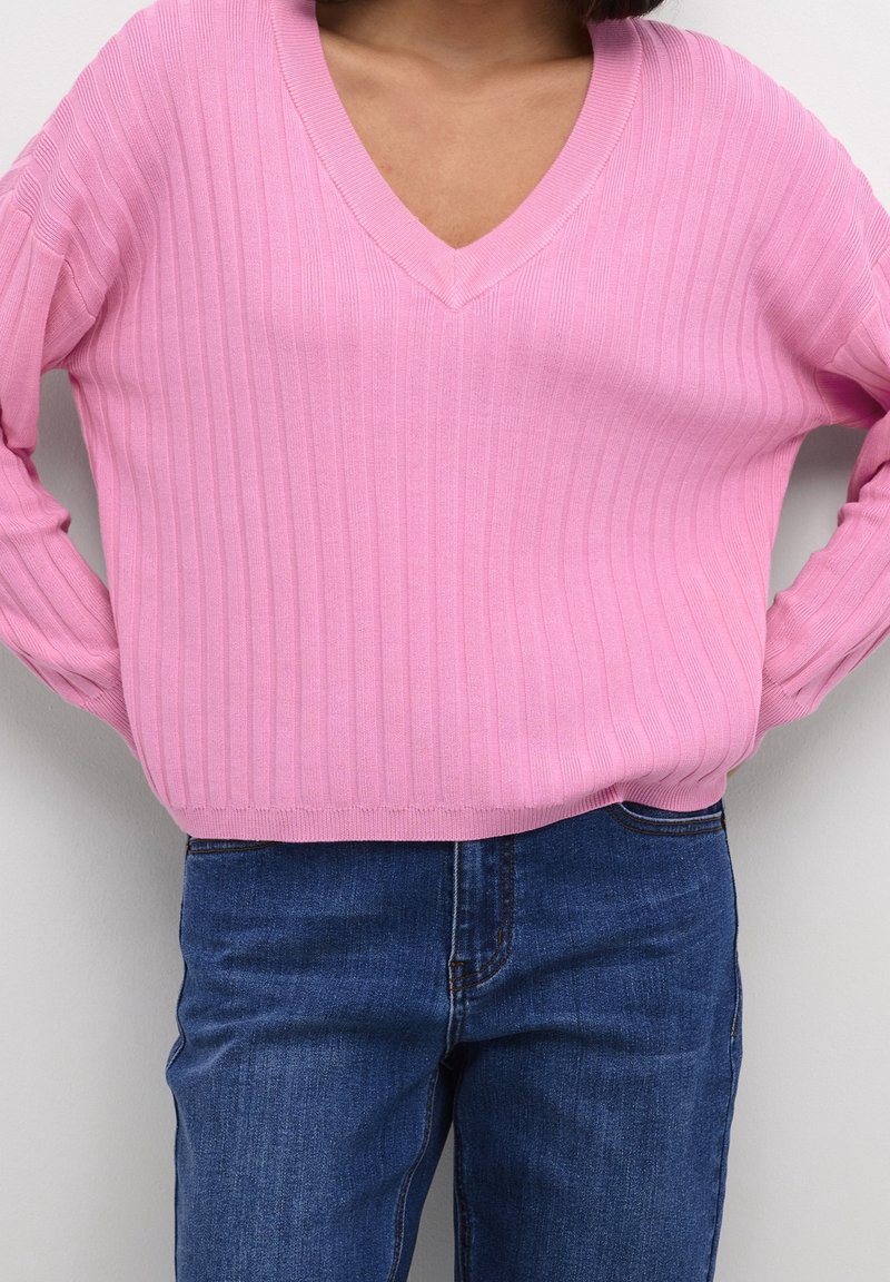 Pull en maille côtelée rose avec un col en V et des manches longues, associé à un jean en denim bleu. Le tissu a une texture lisse.