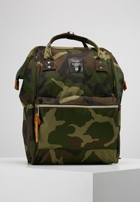 Kamuflaasi-kuvioinen reppu, jossa on ylhäällä kantokahvat, edessä vetoketjullinen tasku ja musta etiketti, jossa lukee "anello", valkoisella pinnalla.