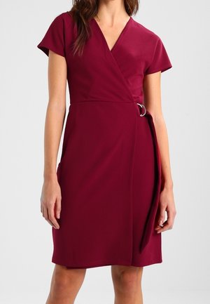 Femme portant une robe portefeuille rouge foncé, à manches courtes et longueur jusqu'aux genoux, avec une ceinture nouée sur le côté et un décolleté en V.