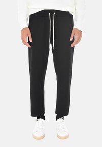 Uomo che indossa pantaloni neri con coulisse, sneakers bianche e un pullover bianco, in piedi contro uno sfondo chiaro e uniforme.