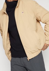 Beige bunda na zip s bočními kapsami, elastickými manžetami a volným střihem, přes tmavý žebrovaný svetr. Malé logo na hrudi.