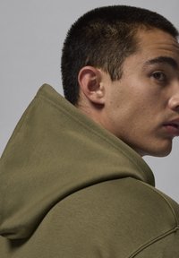 Felpa con cappuccio verde oliva caratterizzata da un tessuto testurizzato, cuciture visibili e una vestibilità rilassata, presentata su uno sfondo neutro.