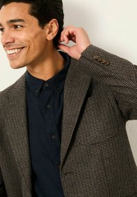 Brauner Hahnentritt-Blazer mit taillierter Passform, Revers und drei braunen Knöpfen an den Ärmeln, kombiniert mit einem dunklen Hemd.