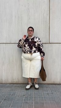 Camisa com estampa de vaca branca, saia branca e sapatos robustos brancos com detalhes em castanho. Bolsa de mão castanha, óculos, encostada a uma parede texturizada.