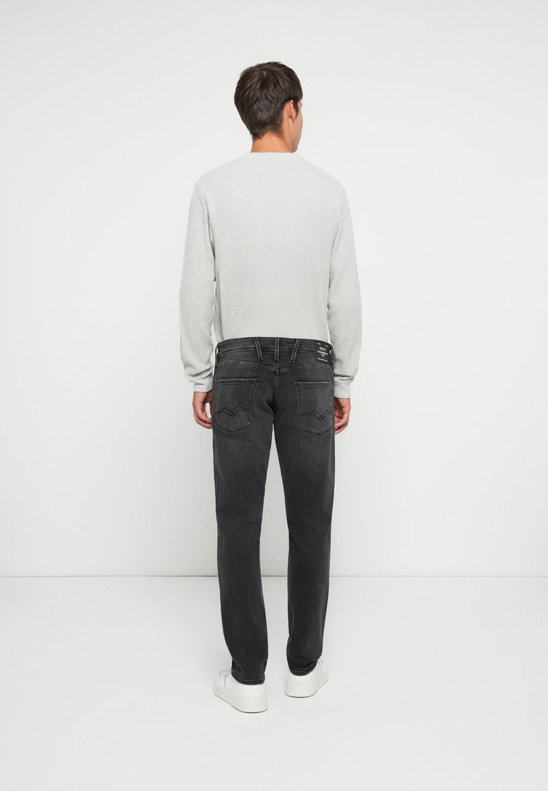 Grijze gebreide trui met lange mouwen, gecombineerd met zwarte jeans. De jeans hebben een achterzakontwerp en worden gedragen met witte sneakers.