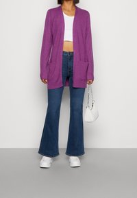 Fialový svetr s dlouhými rukávy, otevřeným předním dílem a dvěma předními kapsami, spárovaný se širokými d žíny a bílými teniskami. Malá bílá kabelka.