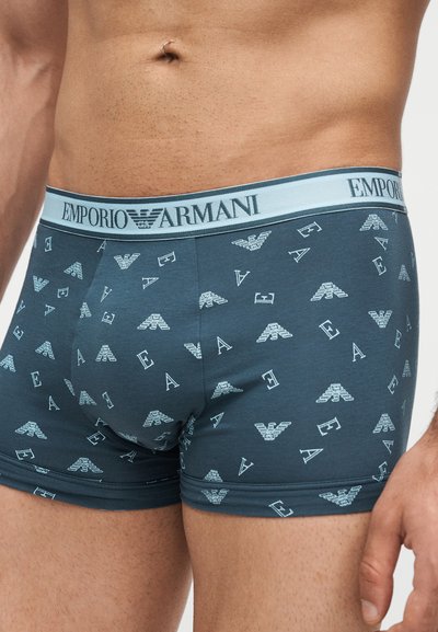 Ανδρικός κορμός που φοράει μπλε boxer Emporio Armani με σχεδιασμένα λογότυπα και φωτεινή γαλάζια ζώνη με επώνυμη σήμανση.