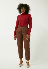 Roter Rollkragenpullover kombiniert mit braunen, gefalteten Hosen. Die Hosen verfügen über einen vorderen Knopfverschluss. Das Model trägt metallic-farbene Absatzschuhe.